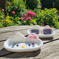 Two handmade white Hello Kitty inspired crochet holders with pink bows in a sunny garden, plain holder in front and sunglasses holder behind